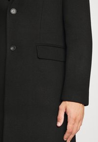 Cappotto di lana nera con una vestibilità sagomata, caratterizzato da due bottoni, una tasca laterale e una superficie liscia. Primo piano della manica e della mano.