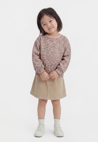 Młoda dziewczyna uśmiechająca się, ubrana w kwiecistą bluzkę z długim rękawem, beżową spódnicę sztruksową, białe skarpetki i białe trampki, stojąca ze splecionymi dłońmi.