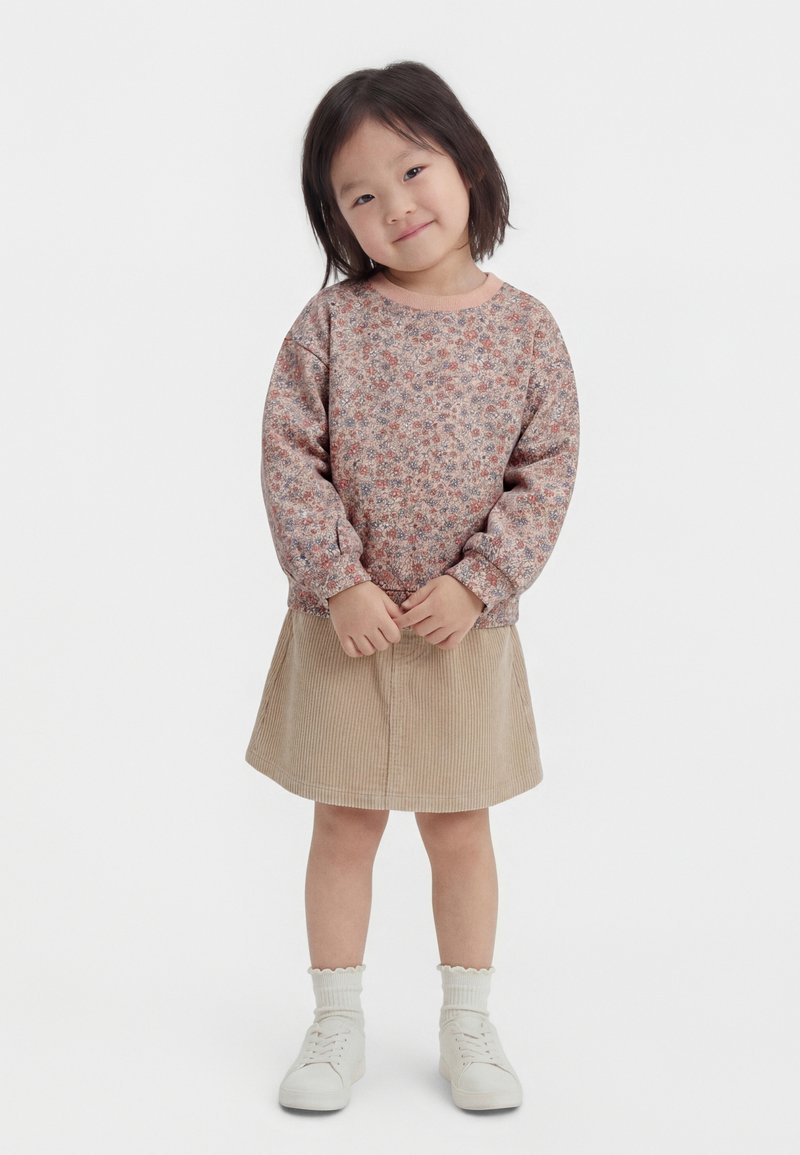 Młoda dziewczyna uśmiechająca się, ubrana w kwiecistą bluzkę z długim rękawem, beżową spódnicę sztruksową, białe skarpetki i białe trampki, stojąca ze splecionymi dłońmi.