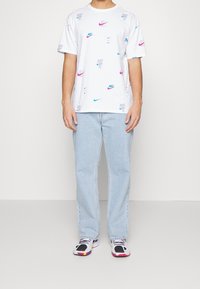 Camiseta blanca de manga corta con logotipos de Nike en azul y rosa dispersos y texto "Just Do It", combinada con vaqueros de color azul claro y zapatillas multicolores.