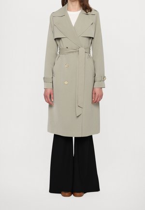 Mujer con un abrigo trench beige con cinturón sobre una camiseta blanca y pantalones anchos negros con zapatos marrones, de pie frente a un fondo liso.