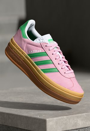 Różowy sneaker Adidas Gazelle z zielonymi paskami i detalem na pięcie, białym językiem i brązową podeszwą, prezentowany na tle szarej betonowej powierzchni.