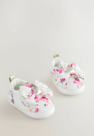Białe buciki dla niemowląt z kwiatowym wzorem w odcieniach różu i zieleni, z dużą kokardą z przodu i metalicznymi akcentami na obcasach, wykonane z miękkiego materiału.