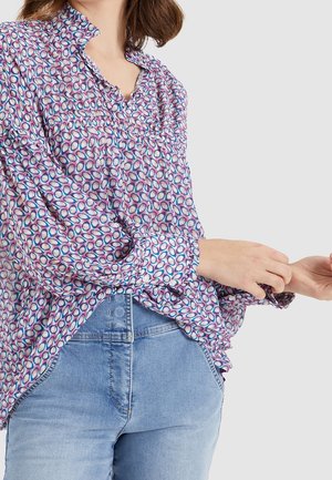 Vrouw draagt een losse blouse met een patroon van blauwe, roze en paarse ovalen, gecombineerd met lichtblauwe hoge taille jeans, terwijl ze haar mouw verstel.