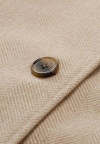 Bruin viergaatjesknop genaaid op beige textuurstof met een visgraatpatroon, waarbij de stofplooi nabij de knop zichtbaar is.