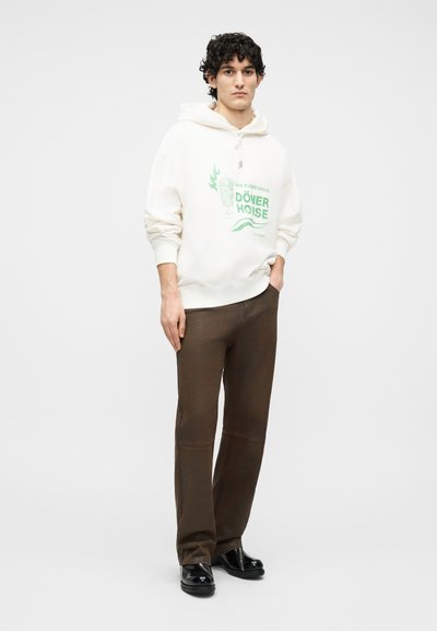 Männliches Model mit lockigem Haar, trägt einen weißen Hoodie mit grünem Text und braune Hose, steht mit einer Hand in der Tasche vor weißem Hintergrund.