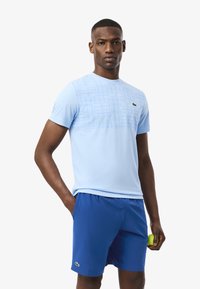 T-shirt azzurro chiaro a maniche corte con motivo a griglia sul davanti, abbinato a pantaloni shorts blu. Presenta un piccolo logo Lacoste sul petto.