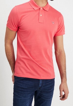 Mężczyzna w koralowej koszulce polo z krótkim rękawem, zapinanej na guziki przy kołnierzyku, oraz ciemnoniebieskich dżinsach, stojący z rękami swobodnie opartymi po bokach.