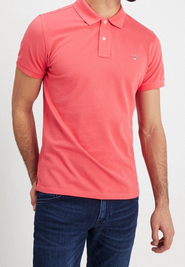 Homme portant un polo corail à manches courtes avec col à boutons et un jean bleu foncé, debout les mains détendues le long du corps.