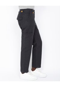 Pantalones cargo negros con múltiples bolsillos, acentos de botones dorados y un ajuste holgado. Combinados con zapatillas blancas sobre un fondo neutro.