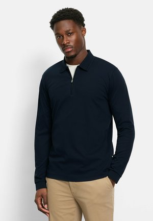Navyblaues Langarm-Polohemd mit Reißverschlusskragen und gerippten Bündchen, aus weichem Stoff gefertigt. Über einem hellen Unterhemd getragen.