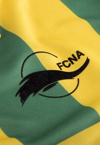Groene en gele sporttricotstof met een zwart geborduurd logo met de tekst "FCNA" en een gestileerd spiraalontwerp.
