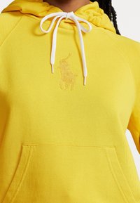 Gul hoodie med snörning i huvan, med en känguruficka och en broderad logotyp i en ljusare nyans. Mjuk tygtextur.