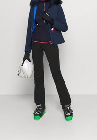 Sneeuwjack in marineblauw met een harige capuchon, zwarte skibroek, handschoenen en groene skibotten. Houdt een witte helm vast met ventilatiegaten.