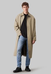 Beżowy płaszcz, luźny krój, długość do kolan, zapięcie na guziki, czarna koszula pod spodem, niebieskie dżinsy i czarne buty, model pozuje na szarym tle.