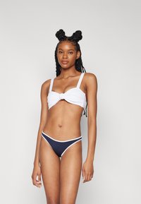 Top bikini bianco con dettaglio annodato, spalline regolabili, abbinato a bikini bottom a vita bassa blu navy con accenti bianchi.