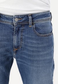 Blue denim jeans feature a classic cut with five pockets, gold stitching, a metal button closure, and a smooth texture.