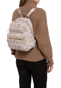 Sac à dos beige avec un design texturé comportant des logos répétés. Il possède une poche zippée à l'avant et des bretelles ajustables, associé à un pull en tricot.