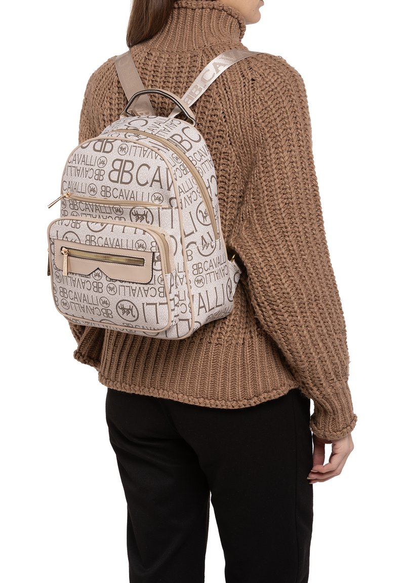 Sac à dos beige avec un design texturé comportant des logos répétés. Il possède une poche zippée à l'avant et des bretelles ajustables, associé à un pull en tricot.