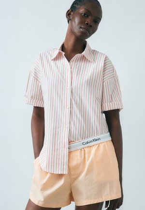 Femme portant une chemise blanche à rayures rouges à manches courtes, boutonnée et rentrée dans un short pêche, avec la ceinture Calvin Klein visible.