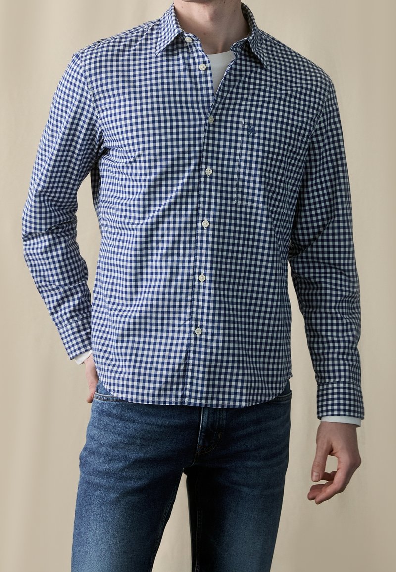 Homme portant une chemise à carreaux bleue et blanche à manches longues par-dessus un maillot blanc, assortie à un jean bleu, debout avec une main derrière le dos.