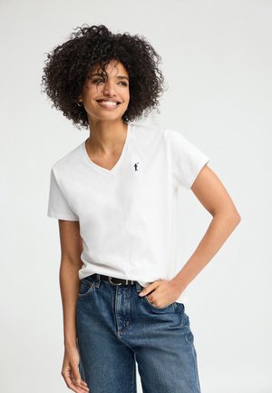 Lächelnde Frau mit lockigem Haar trägt ein weißes V-Ausschnitt-T-Shirt mit kleinem Logo, blaue Jeans und einen schwarzen Gürtel, Hand in der Tasche.