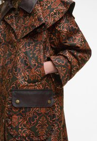 Gesteppten Mantel in Dunkelgrün mit kunstvollen orangefarbenen Blumenmustern, großem Kragen und braunen Lederakzenten an Tasche und Reißverschluss.