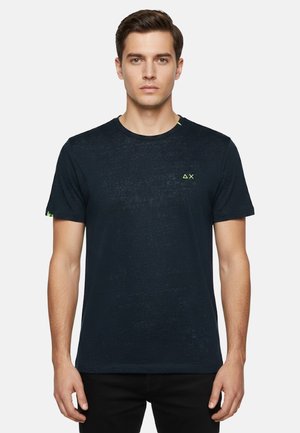 Giovane con capelli corti castani che indossa una maglietta nera aderente con un piccolo logo a triangolo neon giallo e "X" sul petto e sulla manica.