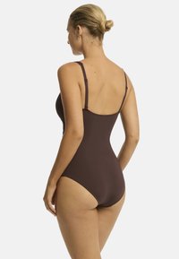 Model trägt einen braunen Einteiler, mit dem Rücken zur Kamera, zeigt das Design des Rückens und die Frisur mit einem Dutt. Weißer Hintergrund.