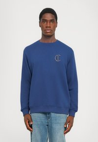 Sudadera azul marino de tejido suave, con cuello redondo y mangas largas. Muestra un pequeño logo bordado en el pecho.