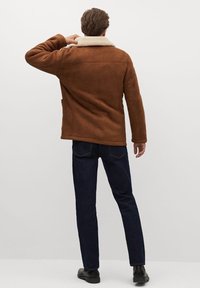 Brun ruskindsjakke med et shearling-krave, der har en lige pasform og to sidelommer, parret med mørkeblå jeans og sorte sko.