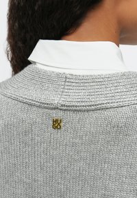 Κοντινό πλάνο του επάνω μέρους της πλάτης ενός ατόμου που φοράει ένα γκρι πλεχτό πουλόβερ πάνω από ένα λευκό πουκάμισο με γιακά, με μια μικρή χρυσή καρφίτσα λογότυπου "HUGO" στο κέντρο, κάτω από τον γιακά.