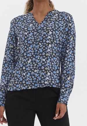 Kvinde iført sort bluse med lange ærmer med blåt, hvidt og gråt blomsterprint, kombineret med sorte bukser.