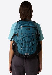 Turkusowy i czarny plecak North Face z regulowanymi paskami, elastycznym sznurkiem bungee, kieszeniami na zamek oraz siateczkowymi kieszeniami bocznymi. Trwała faktura materiału.
