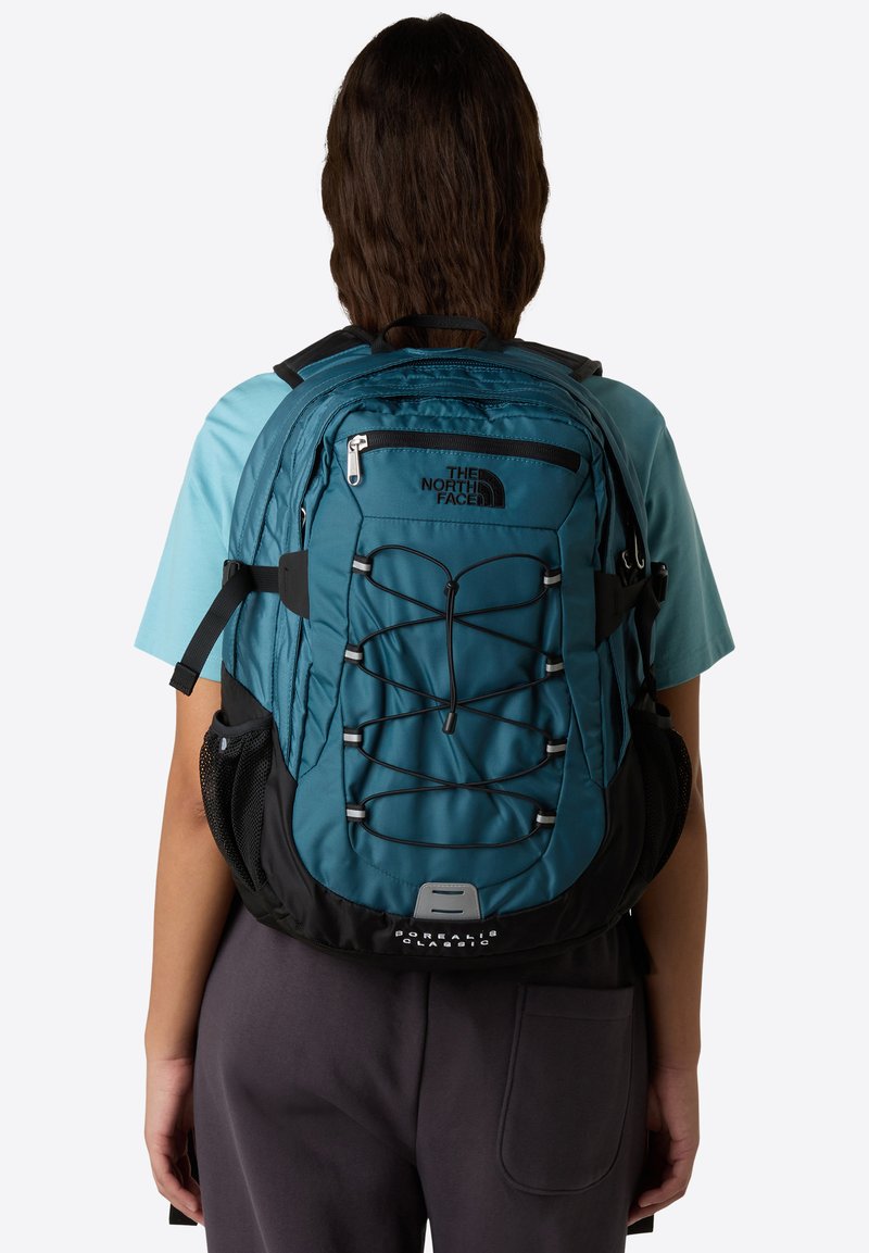 Turkusowy i czarny plecak North Face z regulowanymi paskami, elastycznym sznurkiem bungee, kieszeniami na zamek oraz siateczkowymi kieszeniami bocznymi. Trwała faktura materiału.