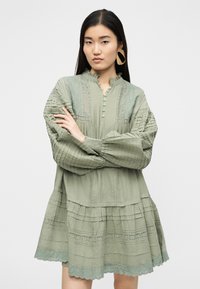 Γυναίκα με μακριά σκούρα μαλλιά σταυρώνει τα χέρια, φοράει ανοιχτό πράσινο φόρεμα με υφή και λεπτομέρειες δαντέλας, μεγάλα χρυσά σκουλαρίκια, ουδέτερο φόντο.