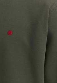 Olive green cotton sweatshirt fabric with a smooth texture, featuring a small red embroidered circular logo on one side.