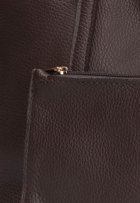 Close-up van donkerbruin gestructureerd leer met zichtbare stiksels en een gedeeltelijk open gouden rits.