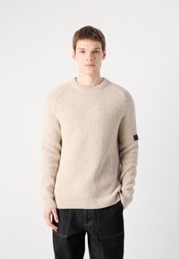 Beige bordázott kötött pulóver, kerek nyakkal, struktúrált anyagból, hosszú ujjakkal. Az egyik bal ujjon egy kis fekete címke található.