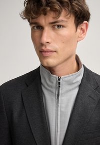 Schwarzer Blazer mit einem taillierten, hellgrauen Reißverschlusskragen. Weiche Textur, maßgeschneiderter Schnitt, minimale Nähdetails. Sauberes, modernes Aussehen.