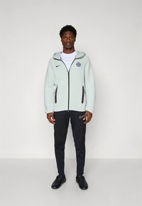 Giacca con cappuccio Nike in pile grigio chiaro con accenti neri, chiusura con zip e logo. Abbinata a pantaloni sportivi neri e calzature nere.