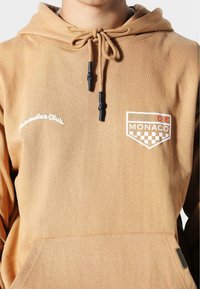 Sudadera beige con cordón y puntas negras, que presenta texto bordado en blanco "Familiar Club" y un parche colorido que dice "Pás de Mónaco."