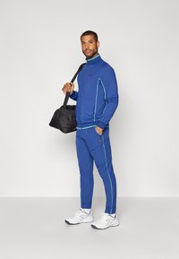 Blauer Sportanzug mit türkisfarbenen Akzenten, bestehend aus einer Reißverschlussjacke und passenden Hosen. Das Modell hält eine schwarze Sporttasche. Weiße Sneakers.