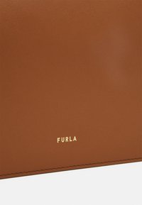 Pruun nahast aksessuaar, millel on sile tekstuur ja õmmeldud servad. Alumisel osal on pronksist "Furla" logo. Minimalistlik disain.