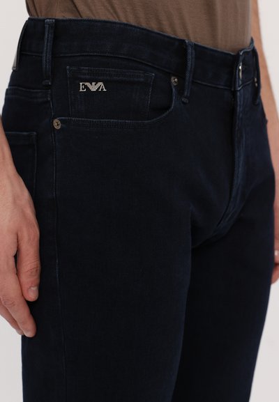 Dark denim jeans, featuring a slim fit, contrasting stitching, and a metal logo accent on the front pocket. Smooth texture and minimal design.