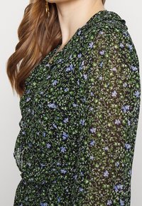 Mujer con largo cabello castaño que lleva una blusa negra transparente con enredaderas verdes y pequeñas flores azules, complementada con un pendiente dorado que cuelga.