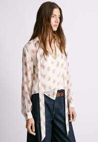 Blusa bianca con motivo floreale in rosa e verde, con chiusura frontale a bottoni, maniche lunghe e orlo inferiore annodato. Abbinata a jeans scuri a vita alta.