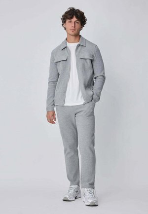 Jeune homme portant une veste zippée gris clair, un pantalon de survêtement assorti, un t-shirt blanc et des baskets blanches, sur un fond clair uni.