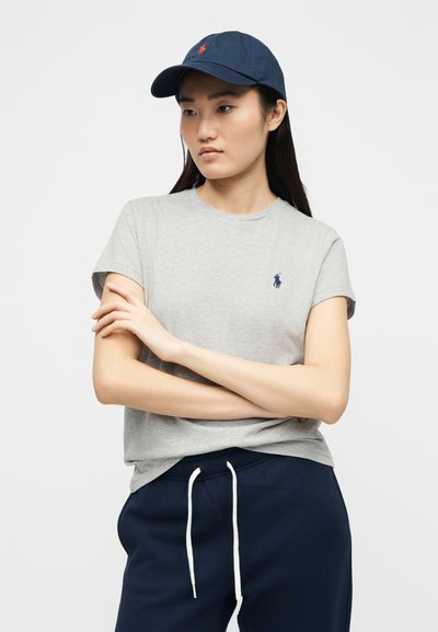 Νεαρή γυναίκα με γκρι μπλουζάκι με λογότυπο, ναυτικά φούτερ παντελόνια με λευκά κορδόνια και ναυτικό καπέλο, στέκεται με σταυρωμένα τα χέρια μπροστά σε ένα απλό φόντο.