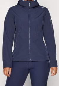 Veste softshell marine avec un col haut, fermeture éclair, deux poches latérales et une poche zippée sur la manche ; texture lisse et coupe ajustée.